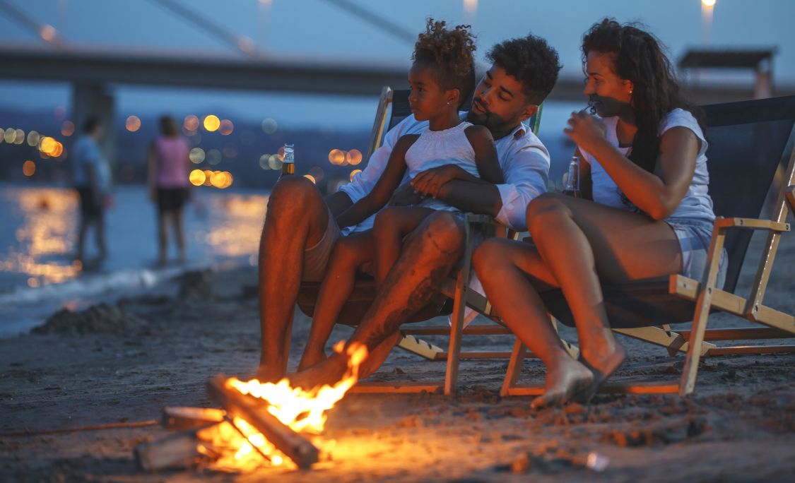A family on the beach at dusk. sitting in chairs by a small campfire