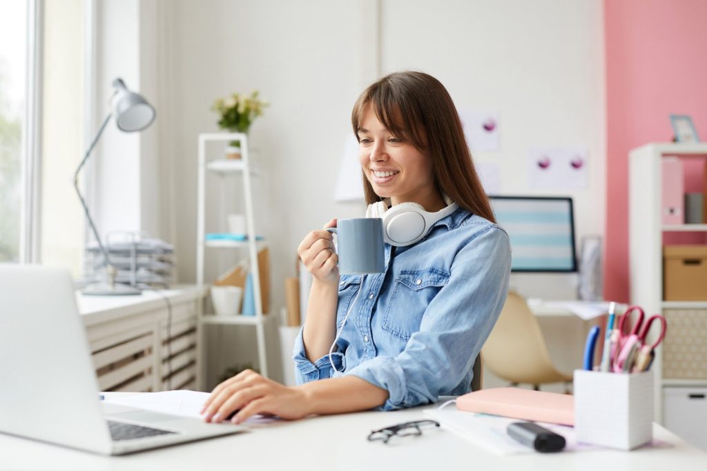 A young woman with a cup of coffee in her hand and her other hand is touching her laptop. She is. smiling as though she is having a meeting at that moment