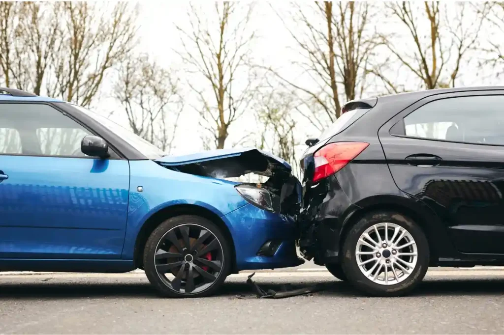 A blue car with a damaged front end collided with the rear of a black car on a street with trees in the background.